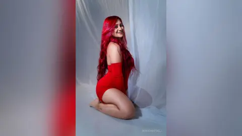 Smiling woman with long red hair wearing a red bodysuit in a studio setting.