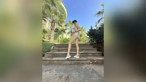 RitaMoonllight stands naked on outdoor stone steps, posing sideways to show her bare buttocks and side profile against a backdrop of palm trees.