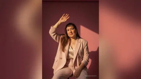 Confident woman in a blush pink blazer and trousers posing against a warm pink background in soft sunlight.