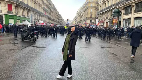 Woman in black coat and green scarf standing on a city street during a crowded outdoor protest.