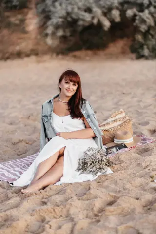 Smiling woman in a white summer dress and denim jacket sitting on a beach blanket during a relaxing sunset.