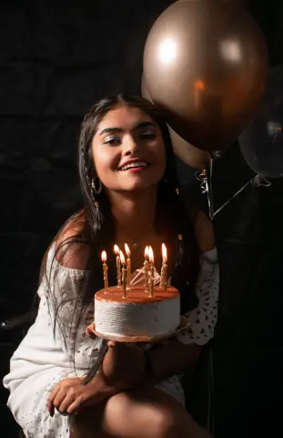Smiling young woman in a white lace dress holding a birthday cake with lit candles near gold balloons.