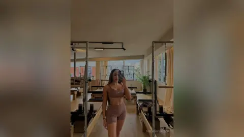 Young woman in brown yoga set taking a mirror selfie in a bright, modern Pilates studio.