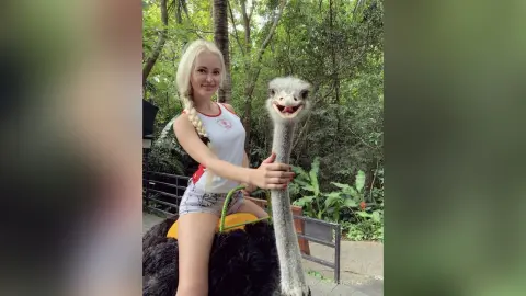 Smiling blonde woman in a white tank top sitting near an ostrich in a lush green garden setting.