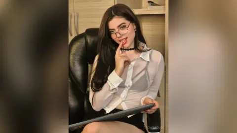 Young woman with long dark hair and glasses wearing a white sheer blouse sitting in an office setting.