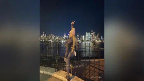 MiaGuerra stands outdoors at night overlooking a lit city skyline, wearing a black puffer jacket, dark leggings, and a pink beanie while making a peace sign.