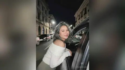 ShaneShaun smiles while leaning against a car at night, wearing a white off-the-shoulder sweater with her long grey-tinted hair down.
