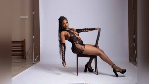 IvyBerry poses on a chair wearing a black lace bodysuit and long lace gloves, showing off her long braids and legs.