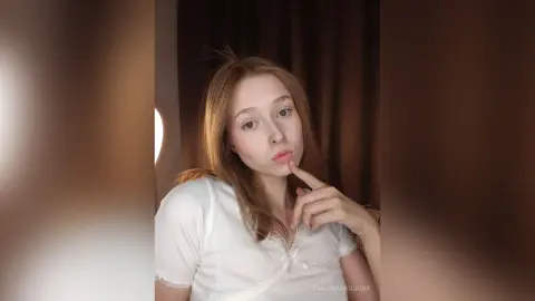 Young woman in a white lace blouse posing thoughtfully in a dimly lit room with soft warm lighting.