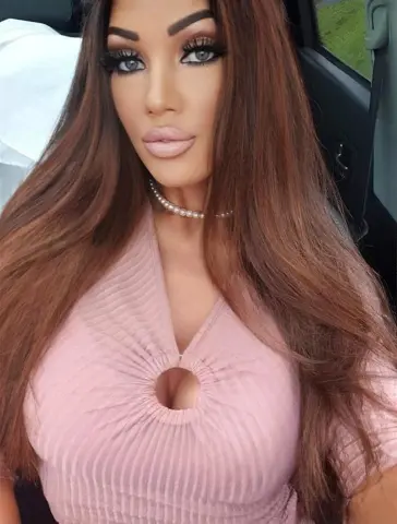 BritishTastyCherryxo with long brown hair wearing a pink ribbed top and pearl necklace while sitting in a car.