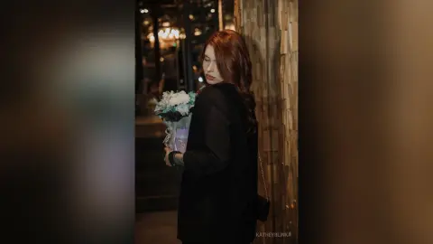 Elegant woman in a black coat holding a bouquet of flowers in a moody, dimly lit urban setting.