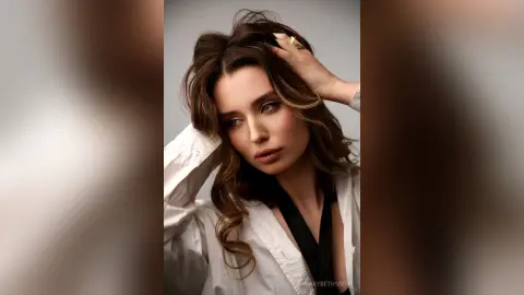 Elegant woman with wavy brown hair wearing a white button-down shirt in a soft, moody studio portrait.