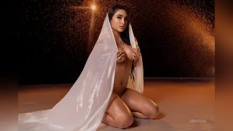 Glamorous woman draped in a white silk fabric posing in a warm, moody setting with golden lighting.