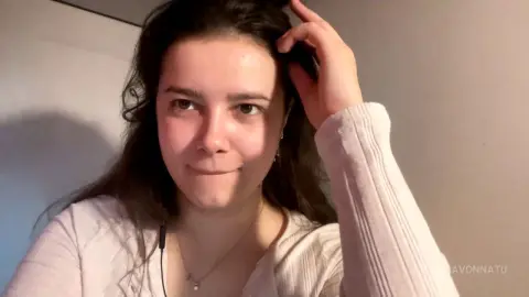 Young woman with long wavy hair wearing a white ribbed sweater in a soft, natural indoor setting.