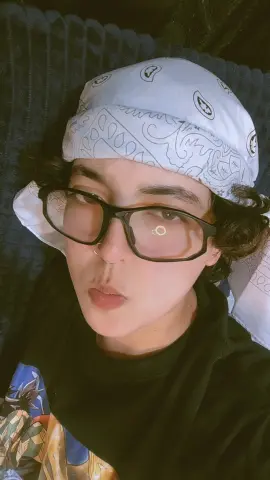 Young person with glasses and a white patterned bandana wearing a black graphic t-shirt in a cozy indoor setting.