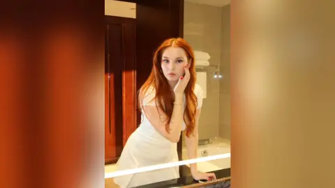 VetaFerr is a redhead in a white dress posing in a bathroom mirror.
