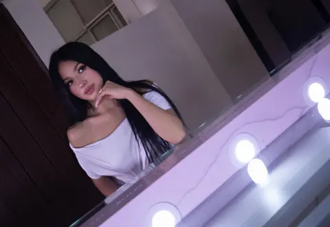 Young woman in a white off-the-shoulder top posing in front of a glowing vanity mirror with a soft mood.