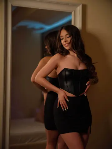 Young woman in a black leather corset and mini skirt posing in a mirror for a moody indoor portrait.