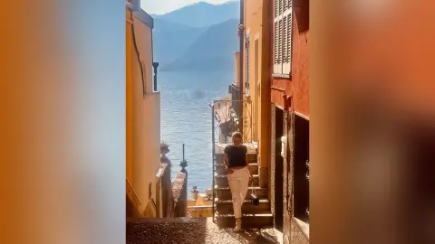 Stylish woman in a black top and white trousers standing on a narrow Italian street overlooking a blue lake.