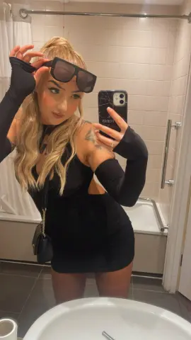 Stylish blonde woman in a black cutout mini dress and sunglasses taking a mirror selfie in a bathroom.