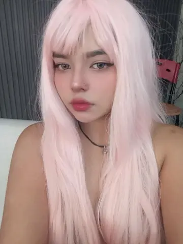Young woman with long pastel pink hair and soft makeup posing in a cozy indoor setting.