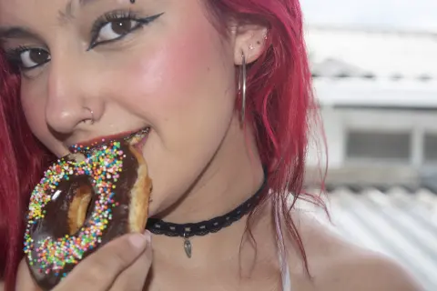 Smiling woman with red hair and black choker eating a chocolate sprinkle donut outdoors in bright sunlight.