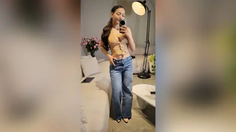 Young woman in a beige crop top and blue jeans taking a mirror selfie in a bright, modern living room.