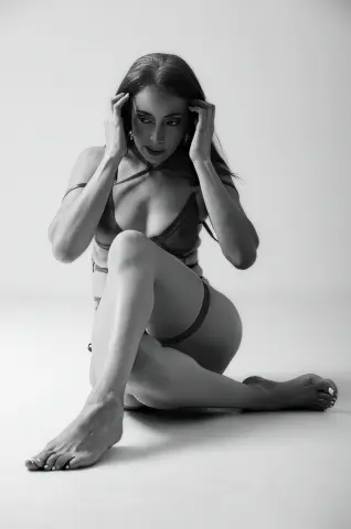 Elegant woman in minimalist lingerie sitting on a white floor in a moody black and white studio portrait.
