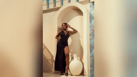 TinaRousell poses in a long black slit dress, leaning against a white stone archway outdoors.