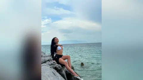 NicoleBella poses naked or partially undressed on grey rocks by the ocean, wearing a black and white bikini top and bottoms.