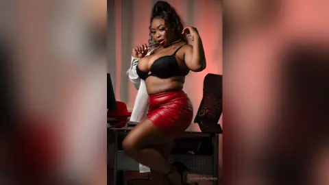 Confident curvy woman in a black bra and red leather skirt posing in a modern office setting.