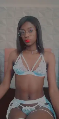 Young Black woman in white lace lingerie posing confidently in a modern bedroom setting.
