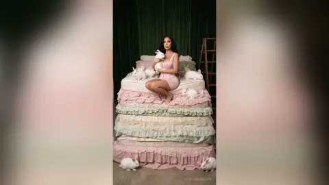 Woman in a pink dress sitting on a tiered ruffled bed surrounded by white rabbits in a dreamy studio setting.