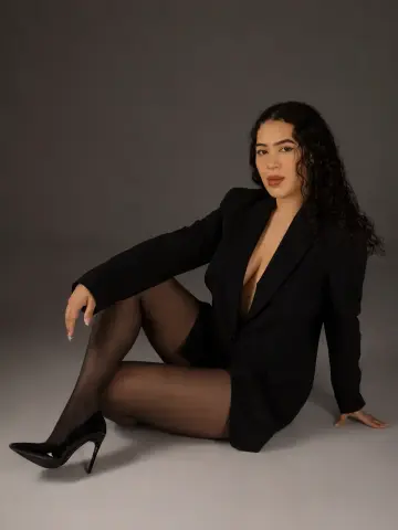 paulinaa_rosee poses in a black blazer and sheer black tights, sitting on the floor with her long, dark curly hair down.
