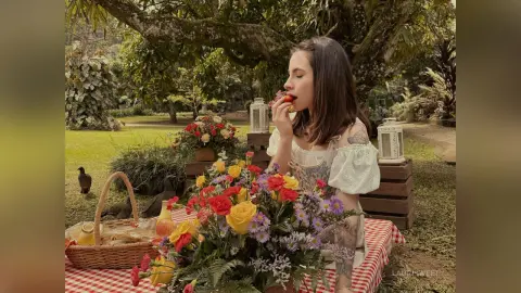 LauriSweet eats a strawberry while sitting behind a large bouquet of colorful flowers at an outdoor table with a red and white checkered tablecloth.