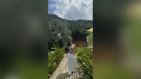 Woman in a white bikini top and flowing trousers standing on a wooden walkway in a lush tropical garden.