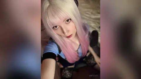 Young woman with long pink and white hair wearing a blue top and black lace outfit in a soft indoor setting.