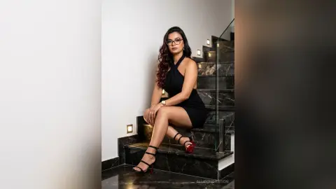 Elegant woman in a black halter dress and strappy heels sitting on modern marble stairs in a luxury home.