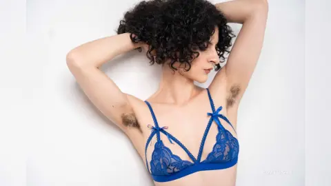 RoxyGraff poses with her hands behind her head, wearing a blue lace bra and showing her dark curly hair and armpit hair against a white background.