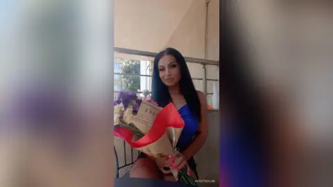 Elegant woman in a blue dress holding a large bouquet of flowers on a sunny balcony.