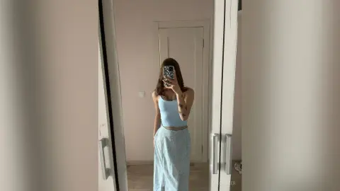 Young woman in a light blue tank top and floral skirt taking a mirror selfie in a minimalist room.