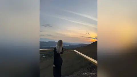 Woman in a black dress looking at a scenic sunset over rolling hills and mountains during golden hour.