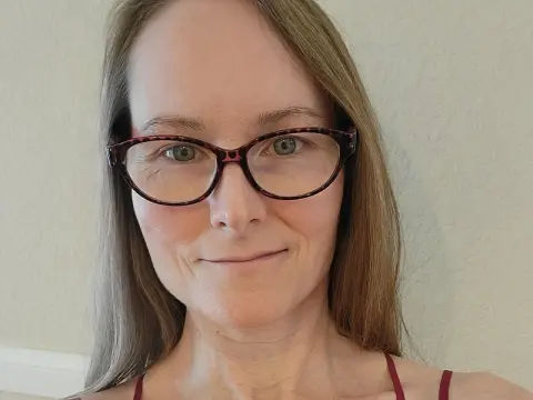 Smiling woman with long hair and tortoiseshell glasses in a bright indoor setting.