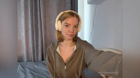 Young woman in a cozy brown hoodie wearing white headphones in a relaxed bedroom setting.