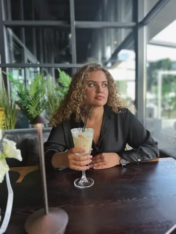Young woman in a black silk blouse sitting at a cafe table with a refreshing iced drink in a bright setting.