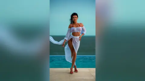 Elegant woman in a white flowing crop top and skirt outfit posing on a balcony overlooking the ocean at sunset.