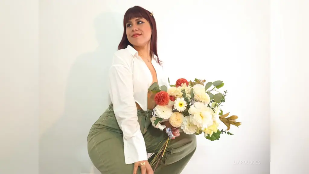 Woman in green denim pants holding a large bouquet of white and pink flowers against a white wall.