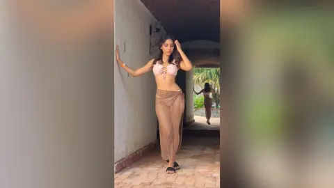 Young woman in a pink bikini top and tan mesh skirt posing in a sunlit Mediterranean hallway.
