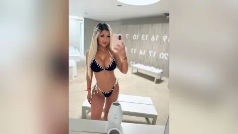 Anita poses in a black string bikini while holding a phone for a mirror selfie in a bright room.