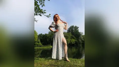 Redheaded woman in a white floral maxi dress posing in a sunny green park by a lake.